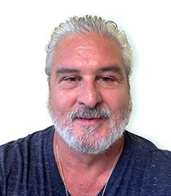 A middle-aged man with white hair and a white beard, wearing a dark V-neck shirt, stands against a plain background. His thoughtful expression suggests he might be contemplating the next innovation in small format printing.