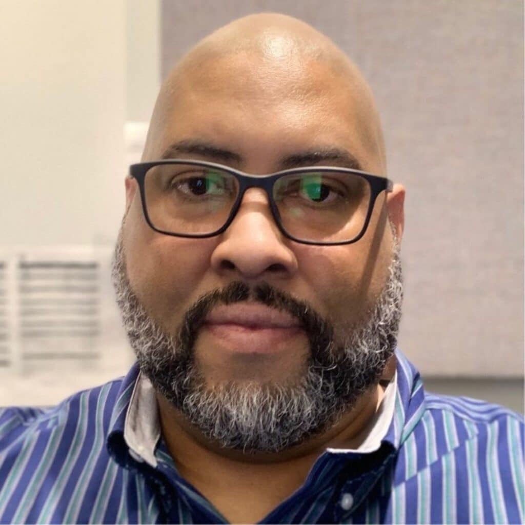 A person with glasses and a beard, wearing a blue striped shirt, is looking at the camera. The background is blurred.