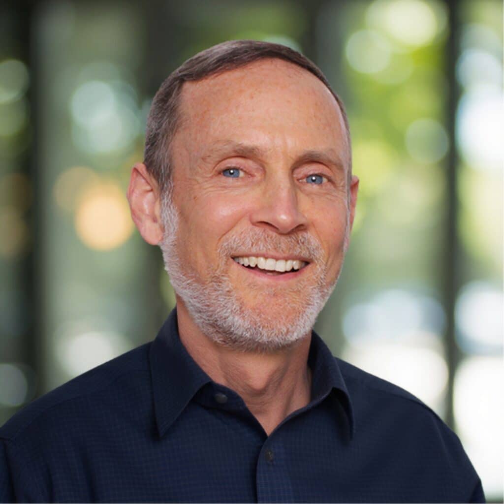 Man with short hair and beard smiling, wearing a dark shirt against a blurred background with green tones.
