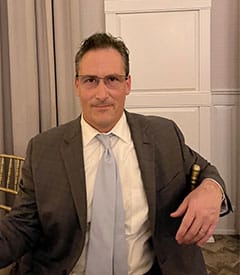 A person wearing a suit, tie, and glasses, seated on a chair indoors with a neutral expression, appears ready to discuss the latest advancements in 3D printing.
