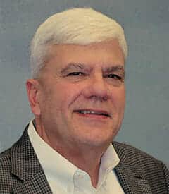 A man with short white hair, wearing a white shirt and checkered blazer, smiles against a plain studio background, his confidence reminiscent of someone ensuring the quality of large format printing.