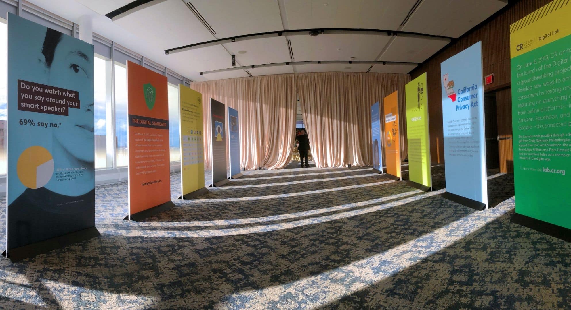 A hallway with a blue carpet is lined on both sides with colorful informational panels, created through large format printing. Drapes are visible at the far end of the hallway.