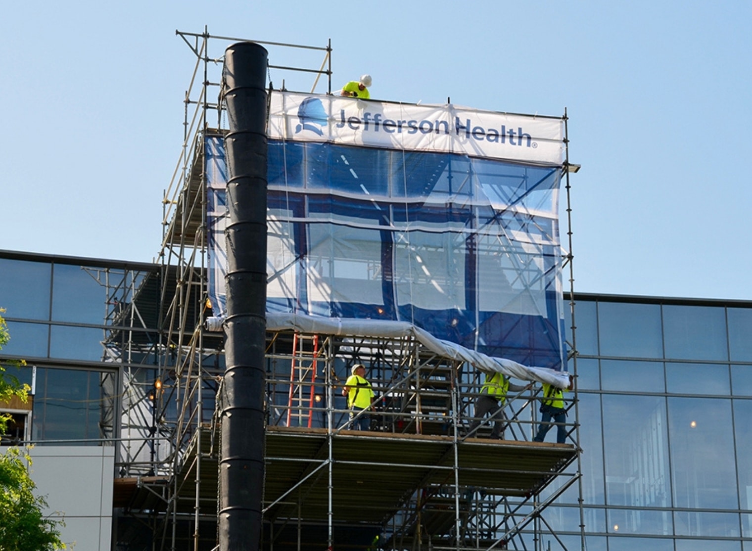 Construction workers on scaffolding in front of a building installing a Jefferson Health sign with precision akin to large format printing.