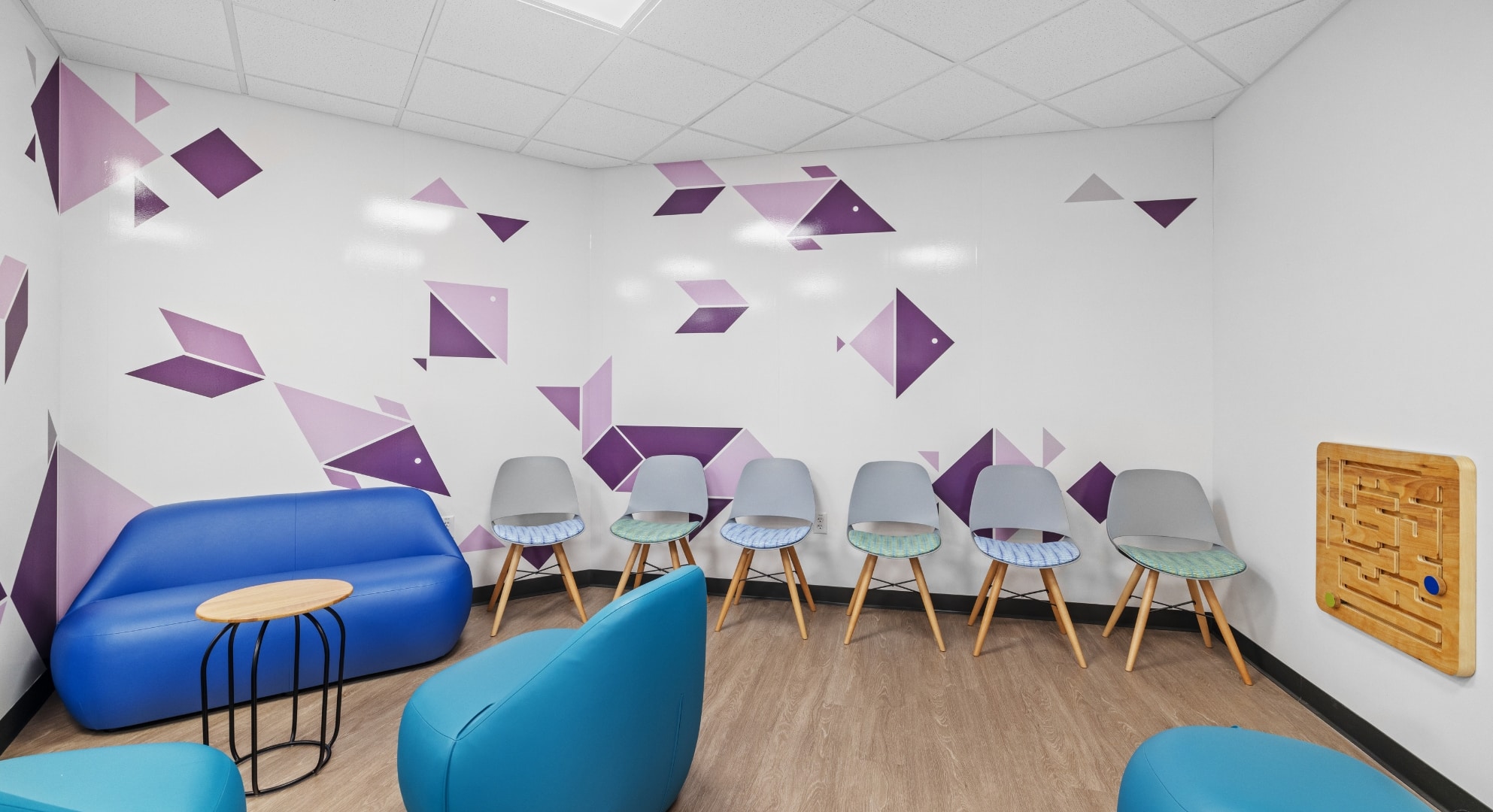 A modern waiting room featuring a blue couch, multiple chairs, geometric purple wall decorations, a small round table, and a wooden labyrinth wall game highlights the innovative integration of 3D printing in interior design.