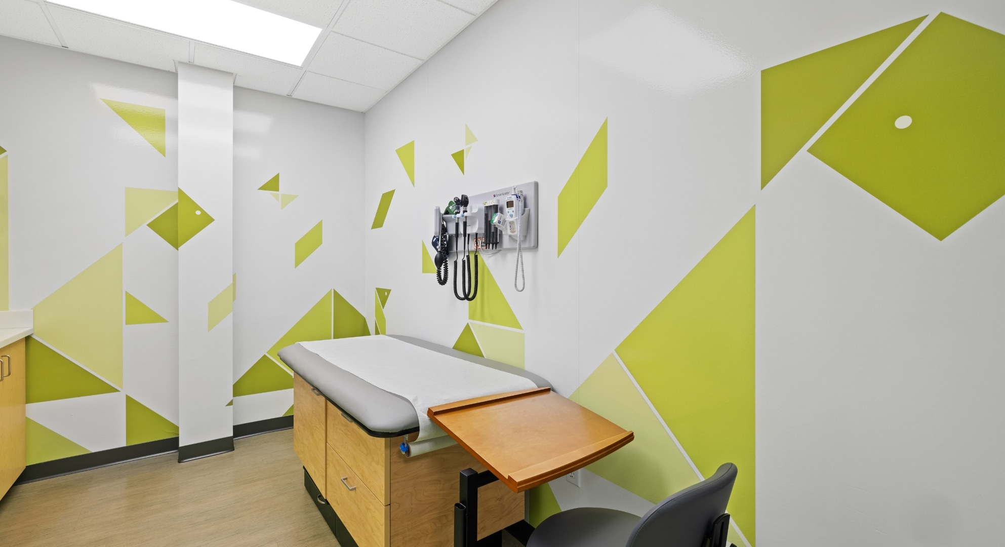 A small, well-lit medical examination room with a cushioned exam table, wall-mounted diagnostic tools, geometric wall art created via 3D printing, and a chair.