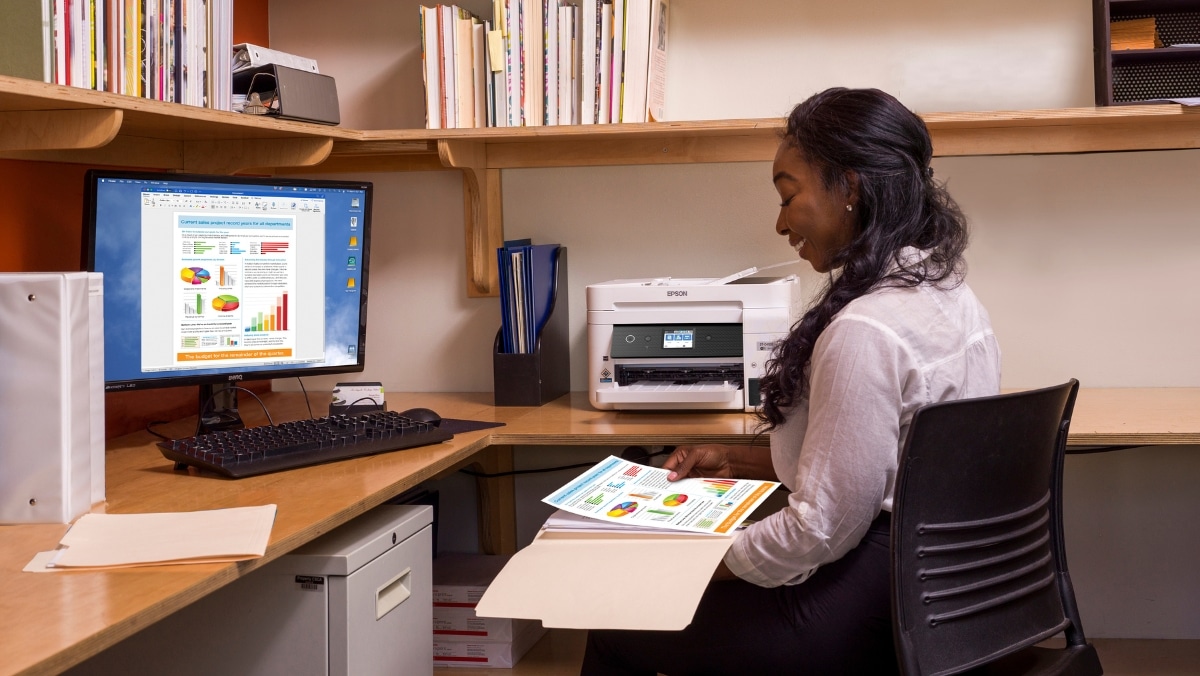 Healthcare 6 - 50_50 A woman sits at a desk holding printed documents with charts, while a computer monitor displays similar graphics. A printer and shelves with books are in the background, hinting at her expertise in 3D printing technology.