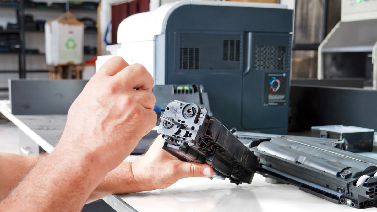 MPS 3 - 50_50 A person is repairing a printer toner cartridge with a tool. A printer and other office equipment are visible in the background, hinting at the versatility of this workspace where both small format printing and large format printing tasks are tackled efficiently.