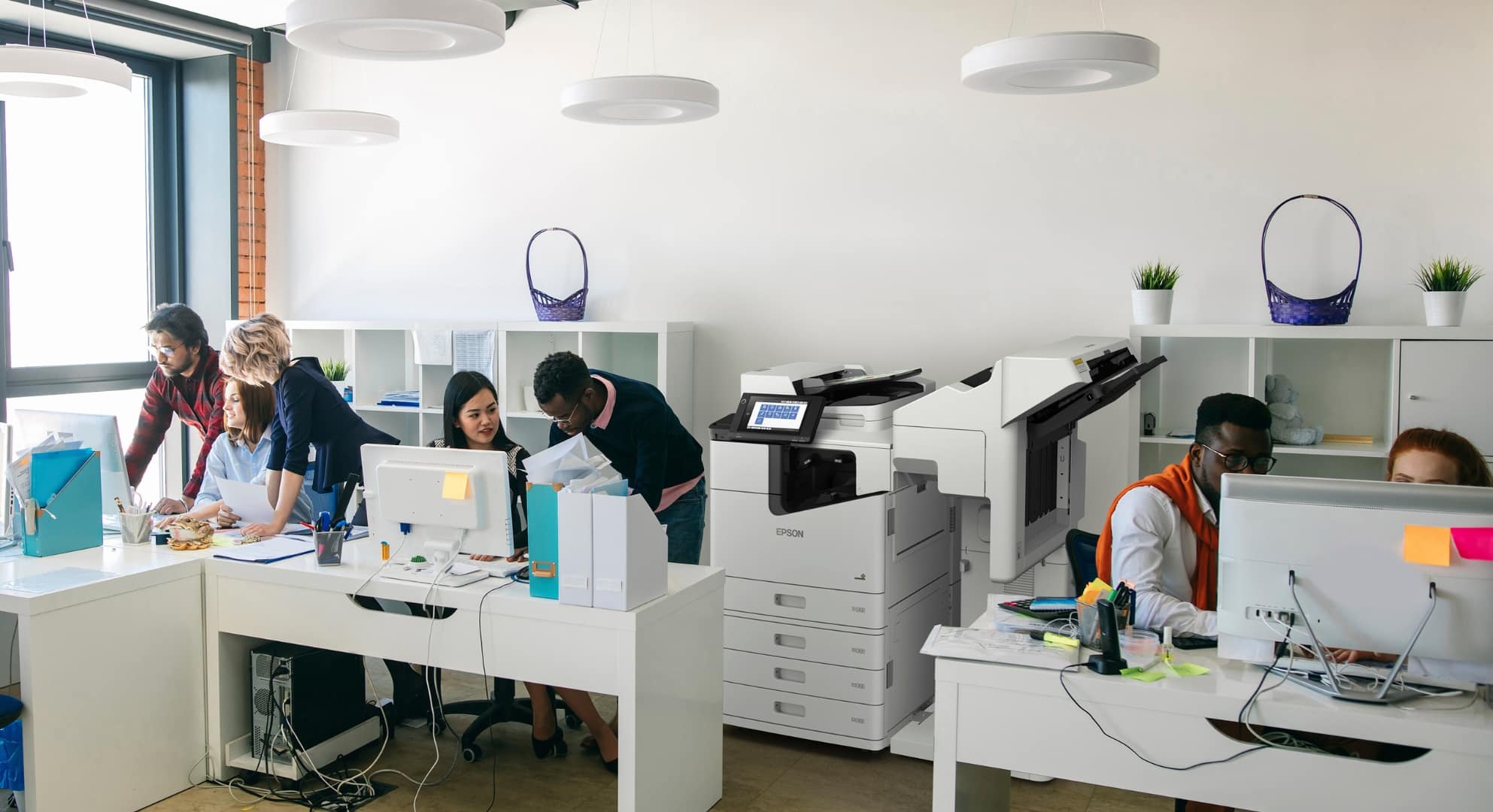 A modern office with several people working at desks equipped with computers and a large printer/copier in the background. Some individuals are collaborating, while others are focused on their tasks, possibly integrating 3D scanning into their workflow.