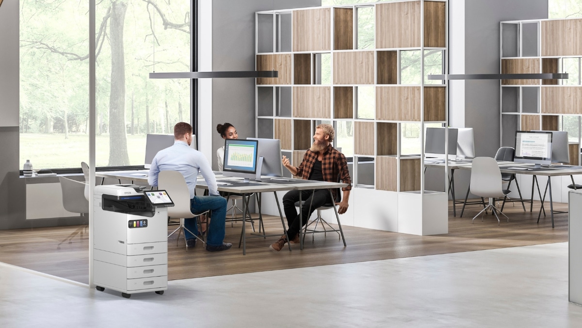 MPS 8 - 50_50 Three people work on computers and discuss something in a modern office with large windows, an open floor plan, and plenty of natural light. To the left, a printer/copier stands ready for tasks like small format printing.