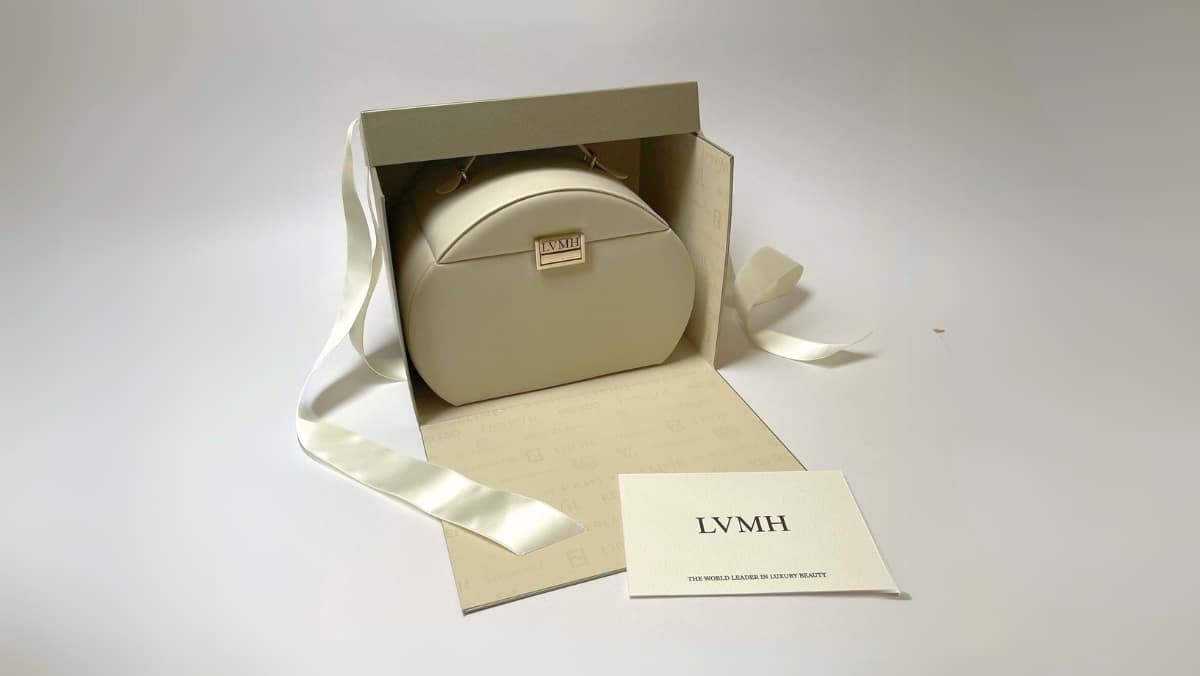 Marketing 1 - 50_50 A round, white designer handbag partially enclosed in a beige box with white ribbons. A white card with "LVMH" and accompanying text, created using small format printing, is placed in front.