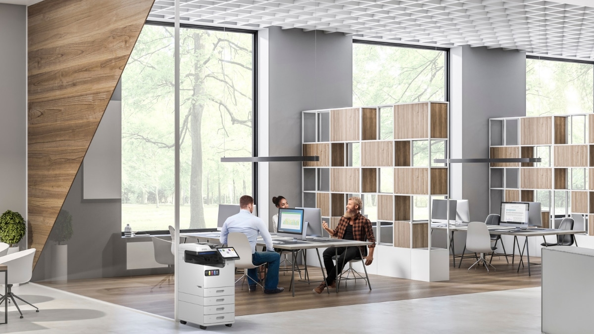Real Estate 5 - 50_50 Modern office with large windows, wooden accents, and white furniture. Three people are collaborating at a table, while another person works at a desk with computer monitors. Trees are visible outside.