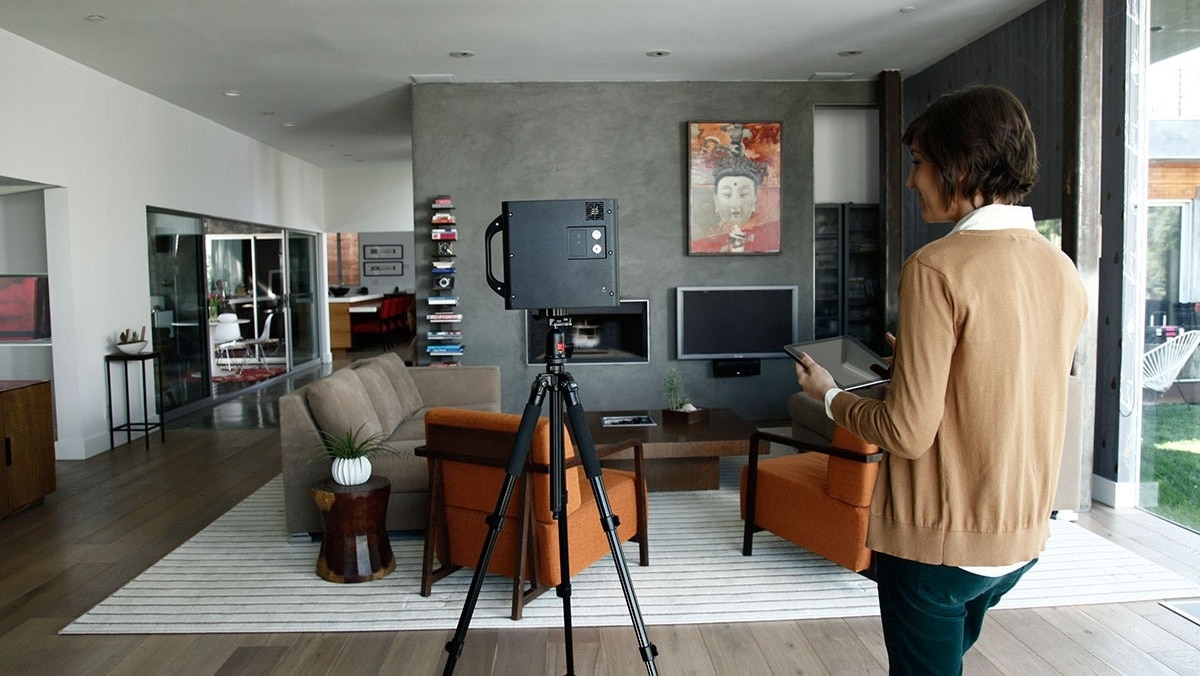 Real Estate - Matterport 3 - 50_50 A person operates a large camera on a tripod in a modern living room with contemporary furniture, a wall-mounted TV, and artwork above it, capturing high-quality images potentially for large format printing.