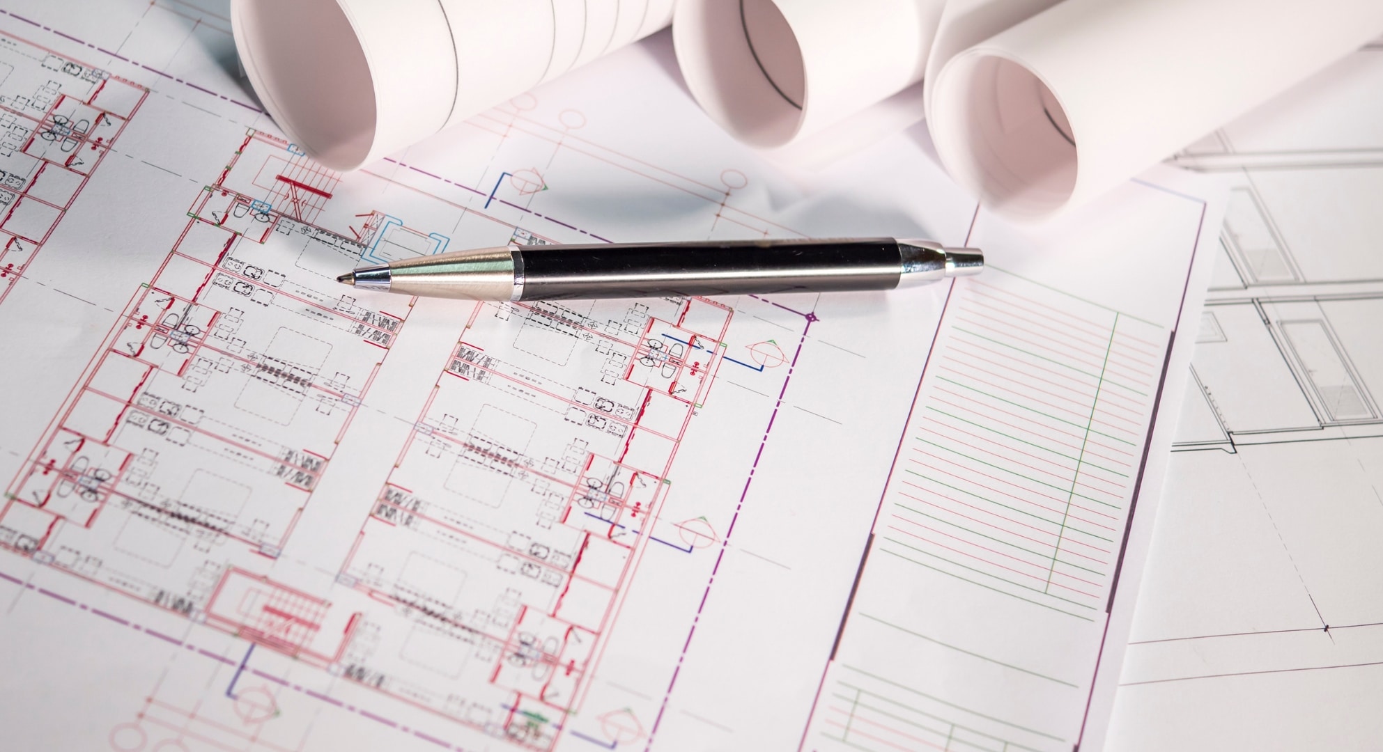 A set of rolled architectural blueprints lie atop a detailed floor plan, with a black pen placed on top, suggesting an ongoing design process that may incorporate 3D printing for prototype models.