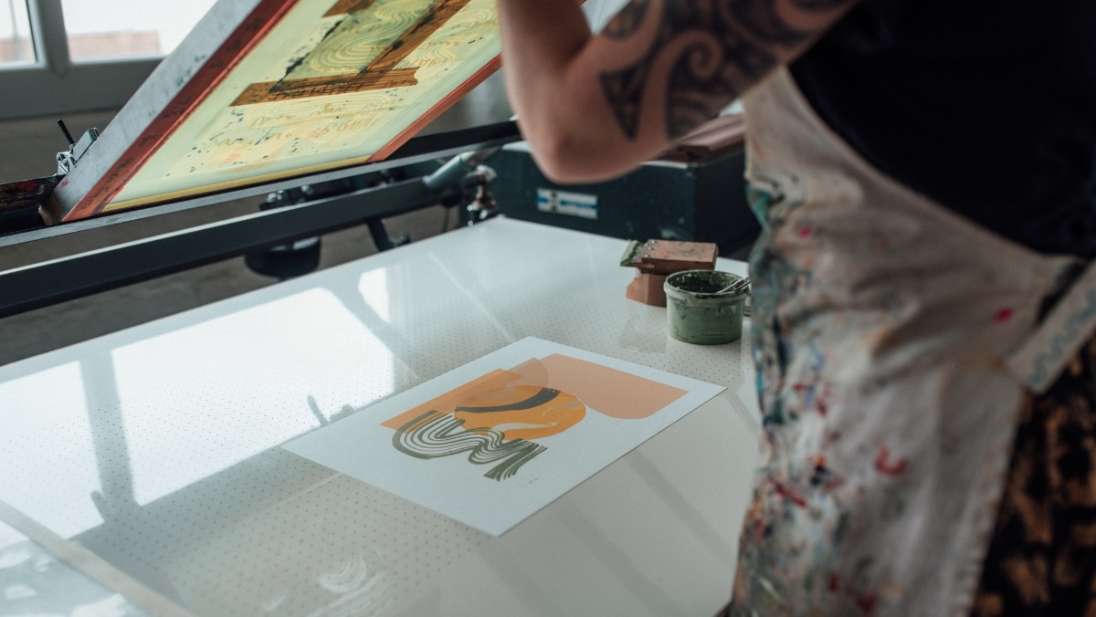 Screen Printing 1 - 50_50 A person wearing an apron and with a tattooed arm is working on a screen printing project, with a sheet of printed artwork and a container of green ink visible on the work surface, blending traditional methods seamlessly in this small format printing setup.