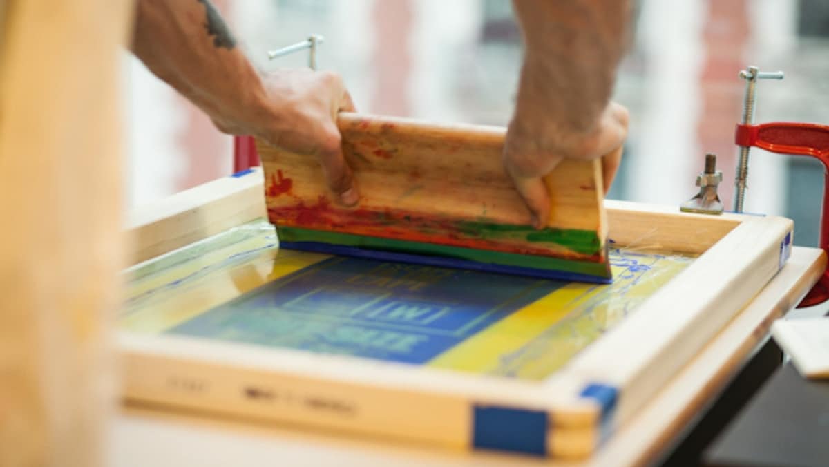 Screen Printing 2 - 50_50 A person efficiently pressing a squeegee over a stencil screen for small format printing, forcing colored ink through the mesh onto a surface below.