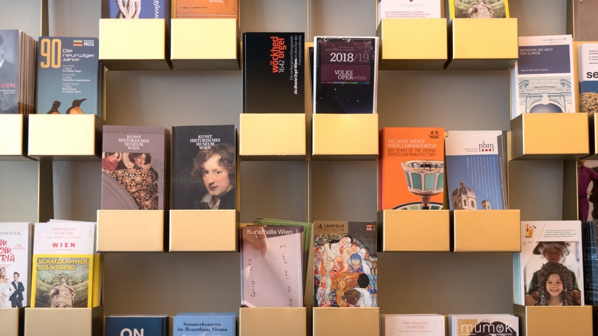 Small Format 2 - 50_50 A wall-mounted display with several gold rectangular shelves holding various booklets, brochures, and postcards, all products of both small format printing and large format printing, arranged in a grid pattern.