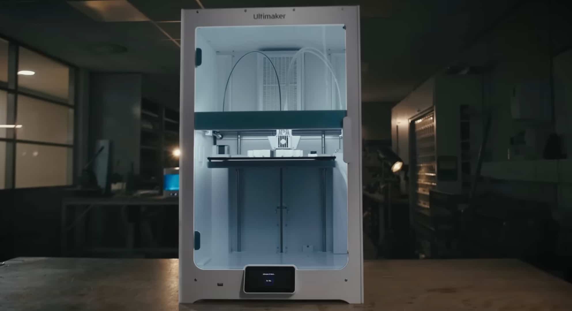 A large Ultimaker 3D printer sits on a workbench in a dimly lit workshop, illuminated from within and ready for use.