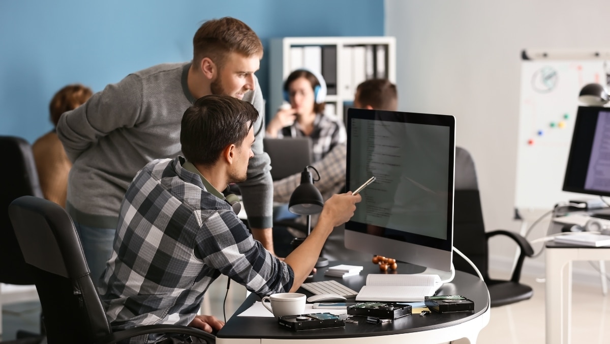 Webstie 50 50 modules Two men in casual attire work together at a computer in an office setting. One is seated, pointing at the screen, while the other leans over. Other people are in the background.