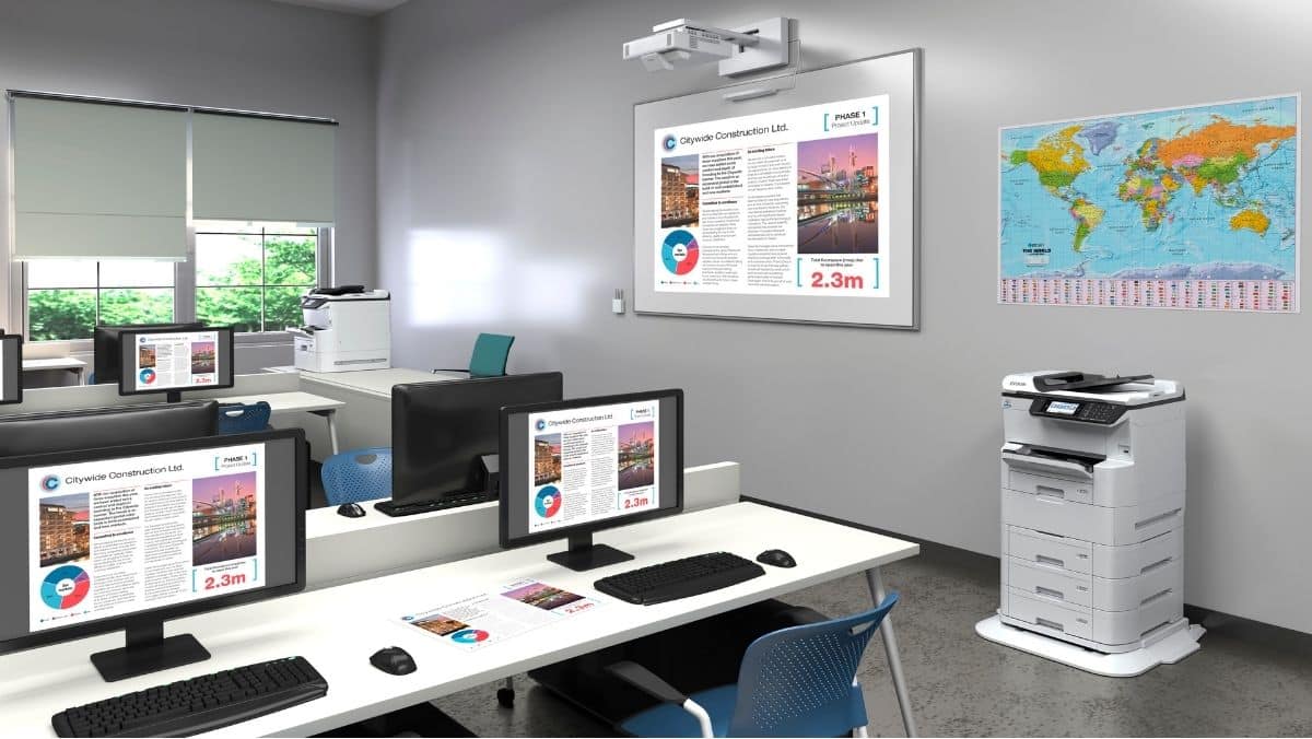 Affordable Modern classroom with computers on desks, a printer in the corner, world map on the wall, and presentation slides displayed on monitors and a wall screen.