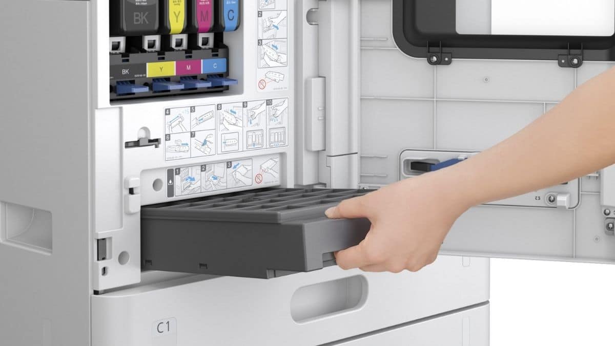 Desktop AM Less Maintenance A hand pulls out a gray printer tray from an open compartment of a color printer, with ink cartridges and instruction labels visible.